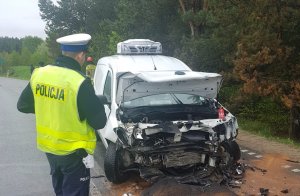 Policjant idzie w stronę rozbitego samochodu, który stoi na drodze