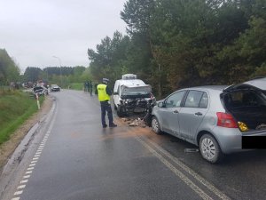 Policjant idzie drogą w kierunku białego rozbitego samochodu stojącego na drodze, za jego plecami stoi inny szary samochód także z rozbitym przodem.