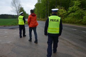 Dwaj policjanci stoją na jezdni w miejscu gdzie doszło do wypadku drogowego, pomiędzy nimi mężczyzna w pomarańczowej kurtce.