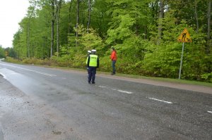 Jeden z policjantów stoi na skraju jezdni po prawej stronie a obok niego w pewnym oddaleniu od siebie swoi drugi policjant oraz mężczyzna w pomarańczowej kurtce - zarządca drogi. Rozmawiają.