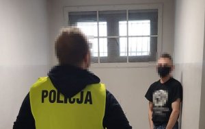 W pomieszczeniu stoi przodem do policjanta mężczyzna w czarnym t-shircie a przed nim stoi policjant w kamizelce odblaskowej z napisem Policja.