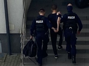 Dwaj policjanci prowadzą zatrzymanego mężczyznę. On ubrany jest w czarne spodnie i czarną bluzę.