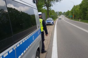 Na pierwszym planie widoczny jest duży radiowóz, tuż za nim na poboczu stoi policjant, a dalej w tle także po lewej stronie drogi stoi drugi policyjny radiowóz.