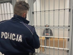 Policjant stoi przed kratą, a za kratą siedzi zatrzymany mężczyzna.