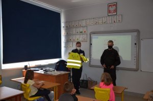 Policjantka stoi przy tablicy, w rękach trzyma kamizelki odblaskowe, które pokazuje uczniom, obok niej stoi strażak w czarnym mundurze.