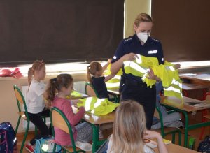 Policjantka chodzi między ławkami i rozdaje uczniom kamizelki odblaskowe.