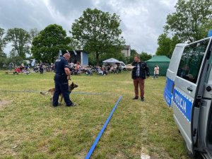 Za radiowozem, policjant z psem na smyczy idzie w kierunku pozoranta, który założył specjalny rękaw ochronny.