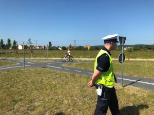 Policjant przechodzi przy miasteczku rowerowym a w tle widoczny jest młody rowerzysta, który zdaje egzamin z jazdy.