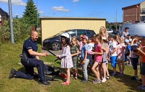 Policjant ukląkł na jedno kolano przed dziećmi, które stoją przed nim w rzędzie, pierwsza dziewczynka przymierza policyjny kask i trzyma pałkę typu tonfa.