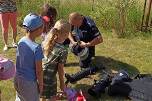 Policjant klęcząc na jednym kolanie przy dzieciach przygotowuje kask ochronny do założenia.