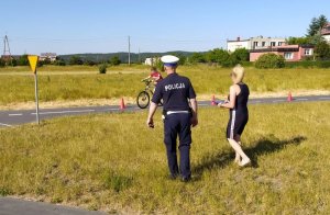 Policjant z nauczycielem stoją na trawie, poza drogami miasteczka rowerowego i egzaminują ucznia.