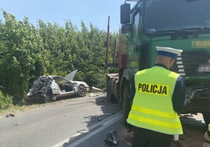 Policjant przygląda się uszkodzeniom na samochodzie ciężarowym. W tle widoczny jest rozbity samochód osobowy