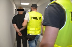 Zatrzymany mężczyzna idzie przodem, na rękach ma zapięte kajdanki, prowadzą go policjanci