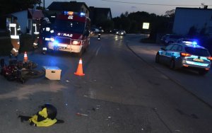 Na zdjęciu na pierwszym planie leży motorower, tuż za nim stoi kilku strażaków, w tle po prawej stronie widoczny jest policyjny radiowóz