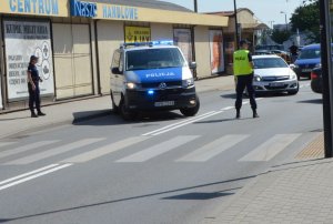 Policyjny radiowóz blokuje ulice, na jednym pasie jezdni stoi policjant, a na chodniku stoi policjantka.