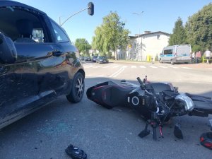 Widoczny jest lewy pok samochodu ford z rozbitą szybą a tuż obok leży, wywrócony na bok motocykl. W tle w oddali widoczny jest radiowóz