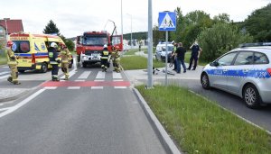 Policyn=jny radiowów stoi na chodniku po prawej stronie, tuż za nim przy drodze dla rowerów widoczny jest przewrócony rower, a za nim w tle wozy strażackie