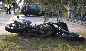 Przewrócony motocykl leży na trawie, w tle za nim na pobliskim parkingu stoi zaparkowany samochód a obok niego po lewej stronie pod wiszącą zabezpieczającą taśmą miejsce zdarzenia, przechodzi policjant.