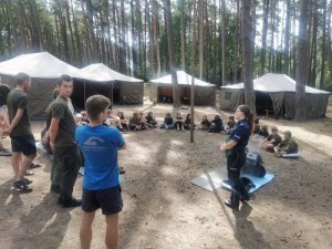 Policjantka stoi  i przemawia do dzieci, które siedzą w kręgu obok i przed nią.