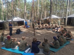 Policjantka stoi  i przemawia do dzieci, które siedzą w kręgu obok i przed nią.