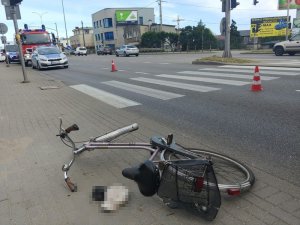 Rower leży na chodniku, a na jezdni dalej stoi radiowóz Policji, straży i znowu za nim Policji