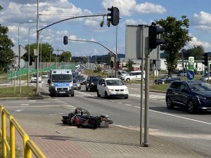 Na przedzie zdjęcia widoczny jest przewrócony motorower leżący na chodniku, kilkanaście metrów dalej stoi policyjny radiowóz na jednym pasie jezdni.