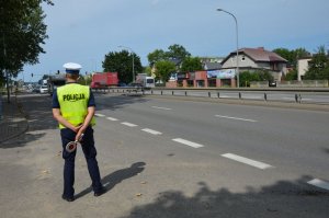 Policjant stoi na poboczu drogi i obserwuje jezdnię, nadjeżdżające samochody, z tyłu za plecami w rękach trzyma biało - czerwoną tarczę do zatrzymywania samochodów