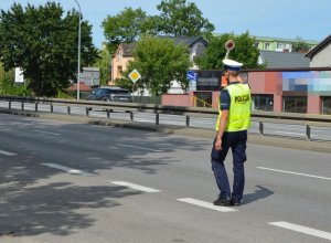 Policjant stoi ina drodze, jedną rękę z tarczą do zatrzymywania pojazdów ma wyciągniętą do góry i daje sygnał do zatrzymania