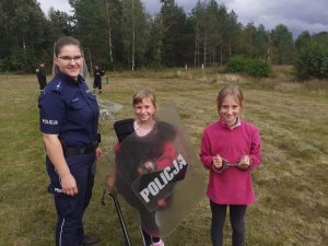 Policjantka stoi z dwójką dziec, które przymierzają elementy umundurowania ochronnego.