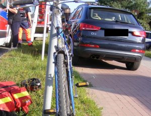 Na pierwszym planie na zdjęciu widoczny jest stojący rower, następnie dalej w tle po prawej stronie widoczny jest samochód audi, a po lewej karetkę pogotowia.