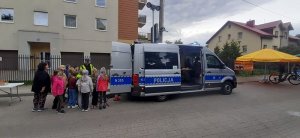 Policyjny radiowóz ma tył otwarty, stoi tam umundurowany policjant oraz przed nim dzieci, które oglądają wyposażenie samochodu. Bardziej widoczny jest bok samochodu.