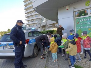 Policjant stoi w czapce, przy radiowozie a obok stoi grupa przedszkolaków z opiekunem.