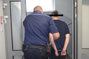 Policjant prowadzi zatrzymanego, przechodzą przez przeszklone z szarym obramowaniem drzwi