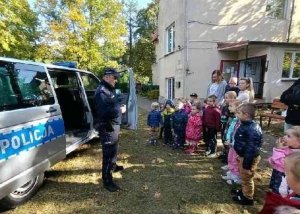 Policjant stoi przodem do przedszkolaków, za plecami stoi policyjny radiowóz