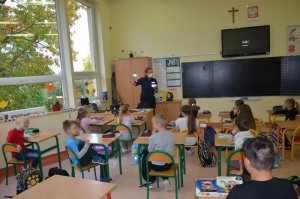 Policjantka pokazuje elementy odblaskowe dzieciom. Stoi za biurkiem, dzieci siedzą w ławkach.