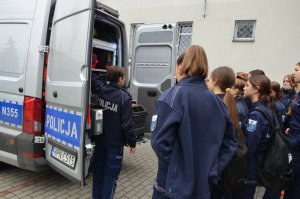 Policjantka prezentuje radiowóz APRD, otwiera tył pojazdu i wyjaśnia młodzieży szkolnej ( w szkolnych mundurkach podobnych do policyjnych), która przygląda się z zaciekawieniem.