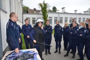 Policjant opowiada, a młodzież  (w szkolnych mundurkach podobnych do policyjnych), przysłuchuje się z zaciekawieniem, Stoją przy stoliku, na którym widoczne są policyjne plakaty.