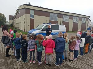 Policjant stoi tyłem do radiowozu, przodem d grupy dzieci, które stoją w półkolu przed nim.