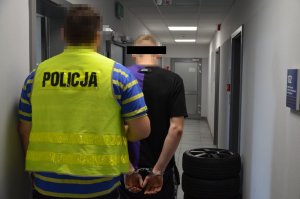 Policjant prowadzi zatrzymanego, w tle po prawej stronie przy ścianie widoczne są odzyskane opony - kompletne koła z alufelgami.