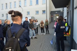 Na pierwszym planie widoczna jest głowa ucznia a przy nim tuż obok stoją inni uczniowie, po prawej stronie stoją policjantki które prowadzą zajęcia z młodzieżą.