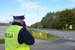 Policjant stoi przy drodze, w dłoni trzyma miernik prędkości trucam, który oparty jest o jego ramię i wykonuje pomiar prędkości. Przyrząd pomiarowy skierowany jest w kierunku nadjeżdżającego pojazdu.