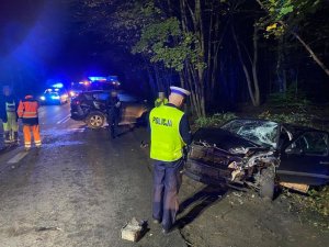 Policjant stoi przed rozbitym samochodem osobowym, w tle widoczny jest drugi samochód, obok policjanta stoi osoba w kamizelce odblaskowej oraz inna osoba ubrana w ciemną kurtkę. W tle za osobami widoczne są samochody służbowe.