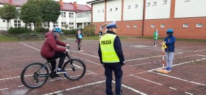 Policjantka na placu - miasteczku rowerowym przeprowadza egzamin. W tle widać dziecko jadące rowerem oraz inne dziec stoją przy ustawionych znakach drogowych.