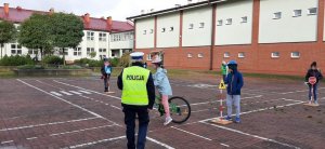 Policjantka na placu - miasteczku rowerowym przeprowadza egzamin. W tle widać dziecko jadące rowerem oraz inne dziec stoją przy ustawionych znakach drogowych.