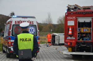 Policjant stoi tyłem do osoby robiącej zdjęcie, za policjantem stoi karetka po lewej oraz wóz straży pożarnej po prawej. Pomiędzy samochodami służb w tle w oddali widać leżący na boku biały samochód.