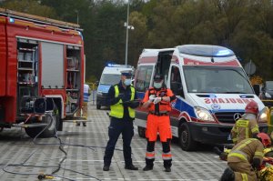 Policjant rozmawia z ratownikiem medycznym, stoją przodem do robiącego zdjęcie, widać że coś uzgadnia, w tle od lewej wóz straży pożarnej, potem za policjantem widoczny jest radiowóz, a potem po prawej widać stojącą karetkę pogotowia.