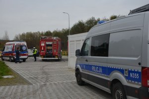 Na pierwszym planie po prawej widać bok policyjnego radiowozu do obsługi wypadków drogowych, dalej w tle widoczna jest karetka i straż pożarna. Przed karetką przy straży pożarnej stoi policjant, który mówi coś do mężczyzny będącego po jego lewej stronie.