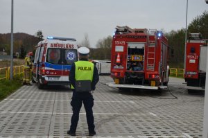 Policjant stoi tyłem do osoby robiącej zdjęcie, za policjantem stoi karetka po lewej oraz wóz straży pożarnej po prawej. Pomiędzy samochodami służb w tle w oddali widać leżący na boku biały samochód.