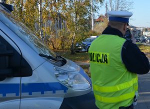 Policjant stoi przy drodze i prowadzi kontrole prędkości, w tle za policjantem widoczny jest fragment radiowozu