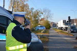 Policjant prowadzi pomiar prędkości, w tle za policjantem stoi policyjny radiowóz wysoki, oraz w pewnym oddaleniu radiowóz osobowy. Ulicą na której odbywa się kontrola jadą samochody.